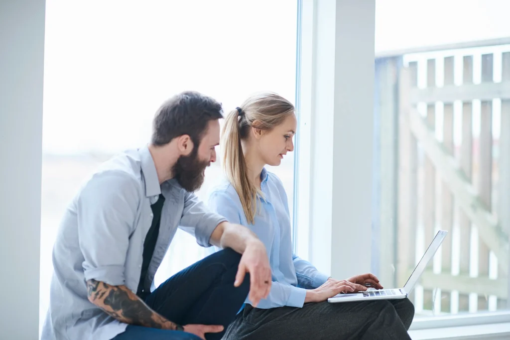 A side-profile shot of two people sitting by a large window in a bright, minimalist room. A woman with a blonde ponytail is typing on a laptop held in her lap, while a man with a dark beard and a tattooed arm sits beside her, looking at the screen.