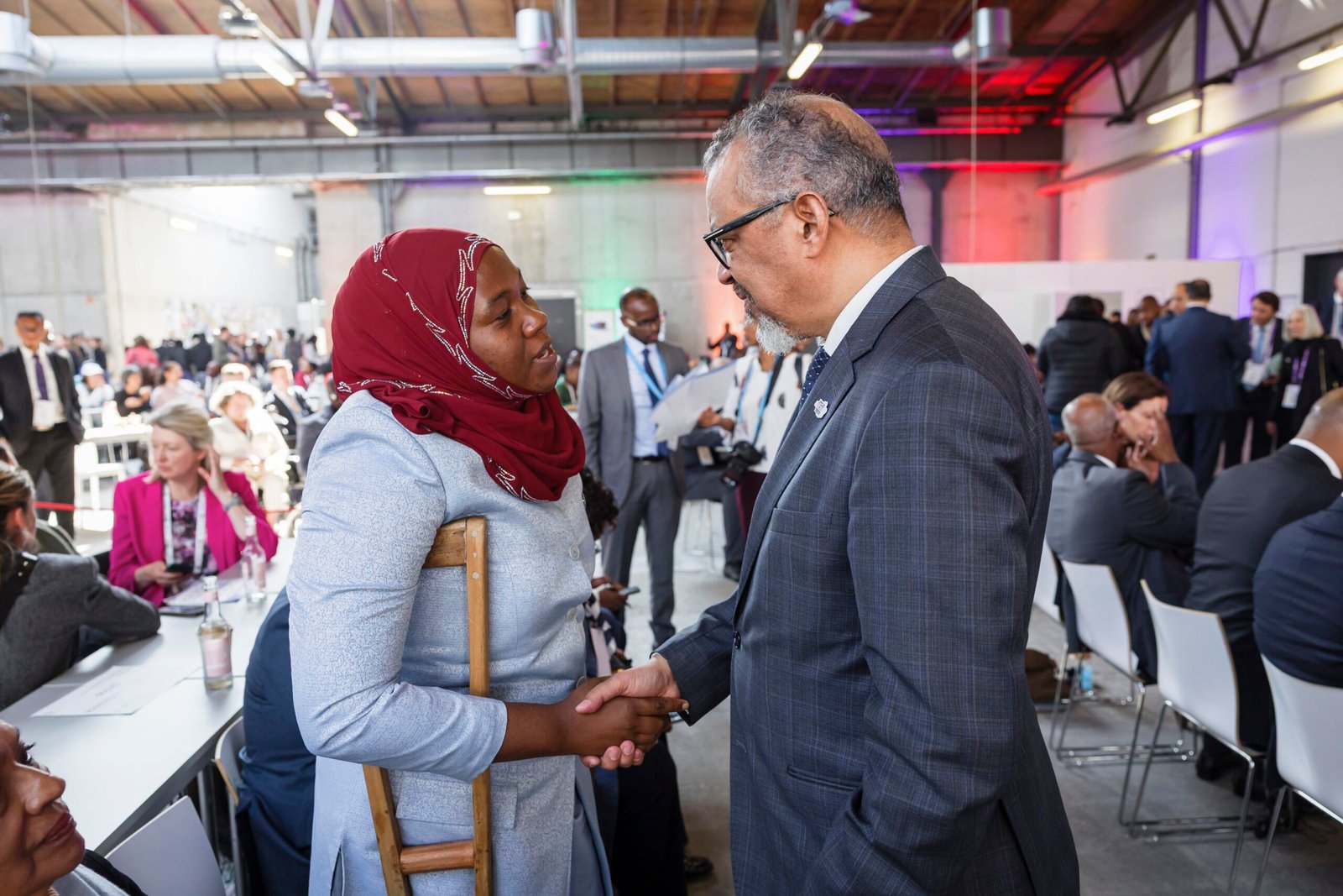 A candid shot of two people shaking hands in a large, busy indoor event space. On the left, a woman wearing a red hijab and a light blue long-sleeved dress uses a wooden crutch for support while shaking hands. On the right, a man in a grey checkered suit and glasses smiles warmly at her. The background is filled with other attendees sitting at long tables under bright, industrial-style ceilings.