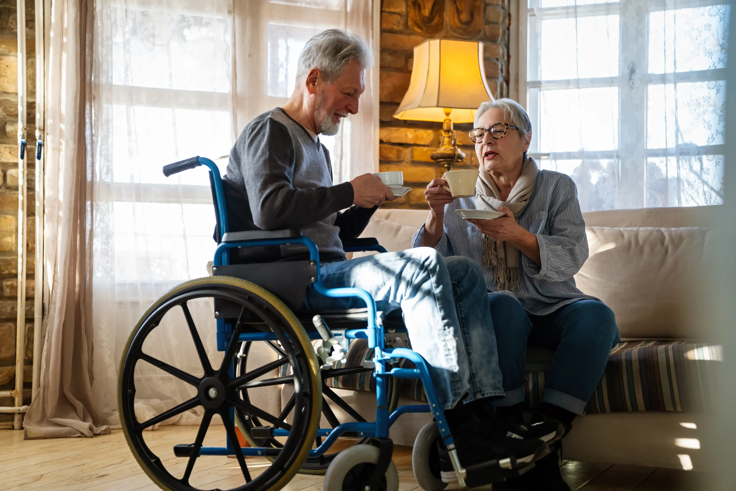 A high-angle, side-profile shot of a smiling senior man with gray hair and a beard sitting in a blue manual wheelchair. He is holding a white teacup and saucer while engaging in a conversation with a senior woman sitting on a nearby sofa. They are in a warm, cozy room with a glowing lamp and large windows in the background.