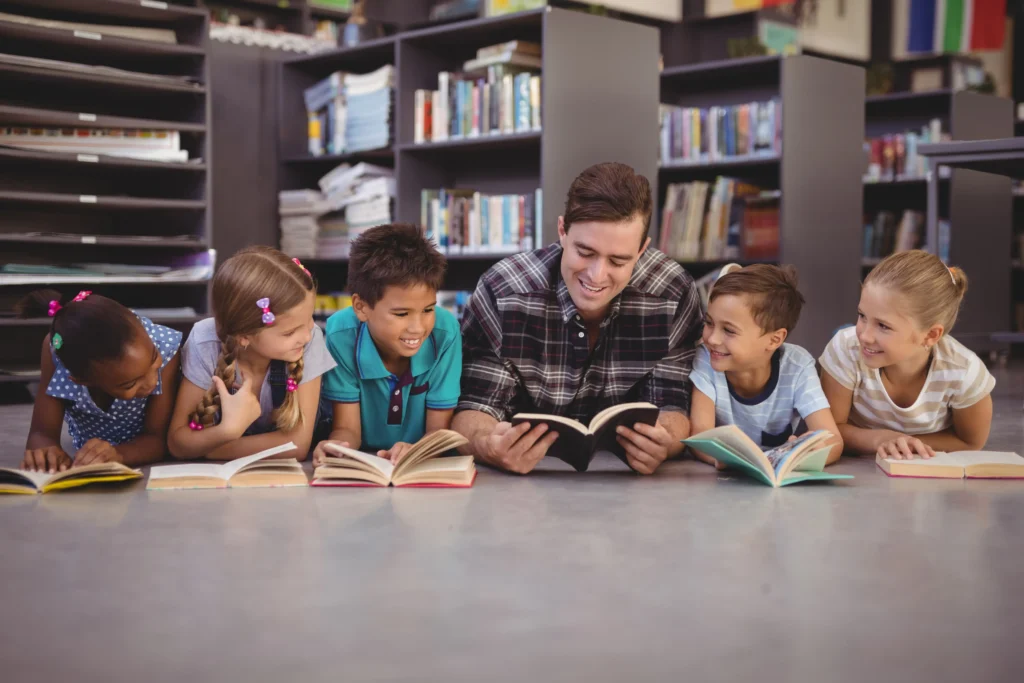 A male teacher and five young school children are lying on their stomachs in a row on a library floor, all holding open books. They are smiling and looking toward the teacher in the center, with bookshelves visible in the background.