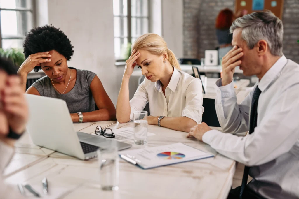 A group of four diverse professionals sitting around a conference table, looking visibly stressed and exhausted. Three of them have their hands to their foreheads or eyes in gestures of frustration or fatigue. Documents, glasses of water, and a laptop are spread across the table.