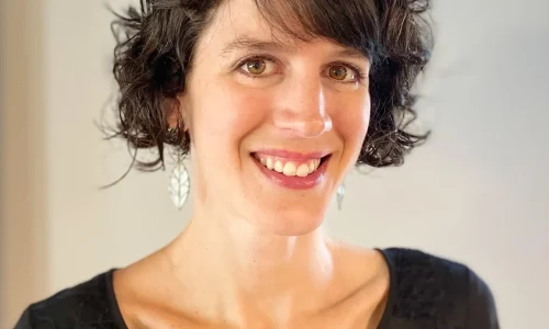 A woman is smiling in this portrait-style photo. She has short, curly dark hair and is wearing a black blouse with lace detailing on the shoulders. She is also wearing silver dangling earrings. The background is plain, emphasizing her friendly and approachable expression. The lighting is soft, giving the image a warm and welcoming feel.